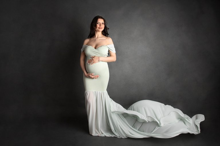 Schwangere, brünette Frau steht vor grauer Leinwand in mintfarbenem, enganliegendem Kleid mit fliegendem Schleier - wunderschöne Indoor Babybauch Fotografie