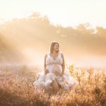 Schwangere Frau steht im Rahmen eines Outdoor Babybauch Shootings mit wunderschönem weißen Wolkenkleid im ersten Sonnenschein am Morgen in nebliger, märchenhafter Kulisse