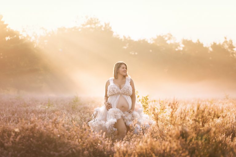 Schwangere Frau steht im Rahmen eines Outdoor Babybauch Shootings mit wunderschönem weißen Wolkenkleid im ersten Sonnenschein am Morgen in nebliger, märchenhafter Kulisse