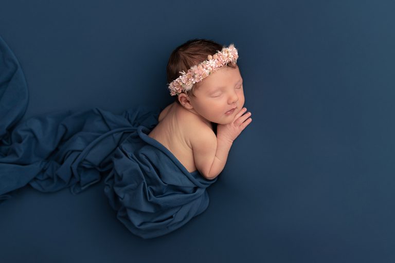 Neugeborenes Baby Mädchen liegt auf einem blauen Tuch auf dem Bauch und trägt einen rosa Haarkranz mit echten getrockneten Blüten – Neugeborenenfotografie für Mannheim, Heidelberg, Darmstadt und Umgebung